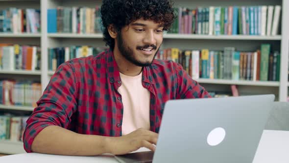 Satisfied Clever Arabian or Indian Student Guy Freelancer Sits in the Library at a Desk with Laptop