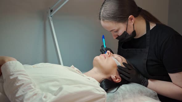 Eyelash Extension Master in a Protective Mask and Gloves Works with a Client Who is Lying on the