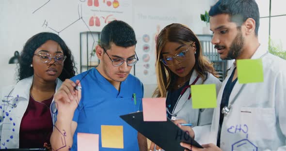 Mixed Race Group of Chemists Standing Near Glass Board and Writing Structural Formulas