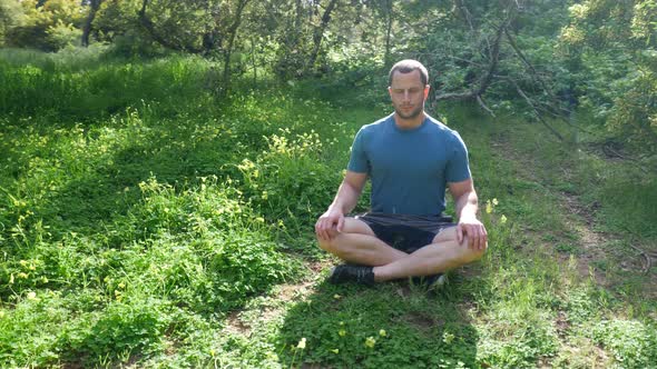 A man sitting in a meditation pose in a green sunny forest meadow practicing deep breathing exercise