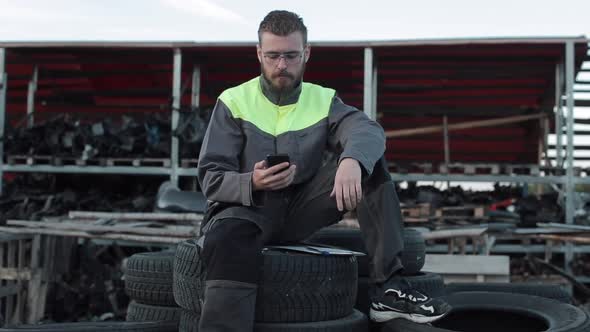 Young Bearded Auto Mechanic with Glasses Sits on a High Pile of Tires and Talks on the Phone. Close