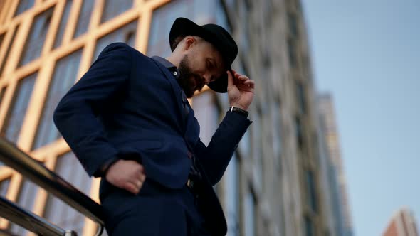 a Bearded and Mustachioed Stylish Man in a Plaid Suit Leans Against the Railing and Adjusts Hat