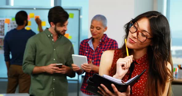 Female executive writing in organizer and colleagues discussing in background