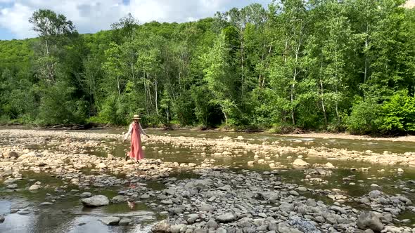 Vintage romantic woman in straw hat, long dress walks on river rocks near forest. Natural scenic