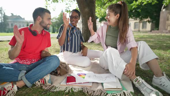 Wide Shot Three Happy Smiling University Students Gesturing Highfive Sitting on Sunny College Yard