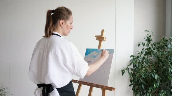 Mature Woman Artist Paints Sky Landscape on Easel Canvas