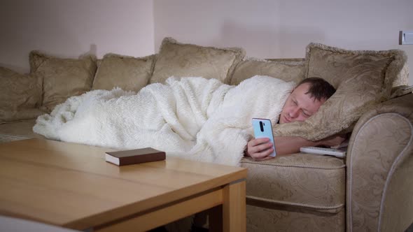 Man Lies on the Couch at Night Watching the TV Holds a Phone in His Hands Evening Lighting