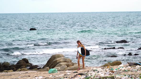 Young surfer in bikini walk in sandal on beach with trash plastic waste and ocean pollution, female