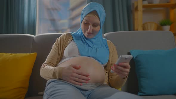 Pregnant Young Muslim Woman in Hijab Scarf Sitting on the Couch Using a Smartphoneinternet Search