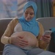 Pregnant Young Muslim Woman in Hijab Scarf Sitting on the Couch Using a Smartphoneinternet Search - VideoHive Item for Sale