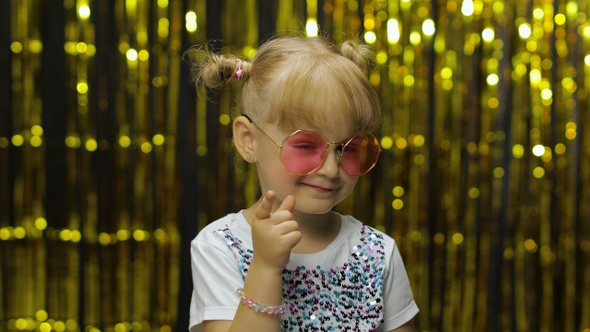 Child Smiling, Pointing Fingers at Camera. Girl Posing on Background with Foil Golden Curtain