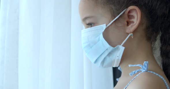 Portrait of a Cute Little Child or Little Girl in a Protective Medical Mask Looks Out the Window