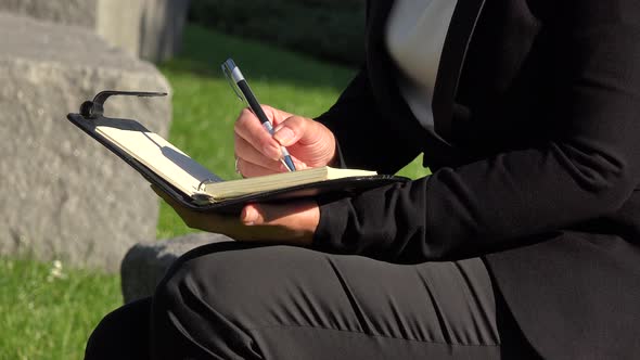 A Middle-aged Woman Sits on a Stone Bench in a Park and Writes Into a Notebook - Closeup