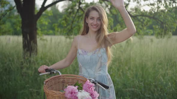Middle Shot Portrait of Charming Smiling Woman Waving Hand As Standing with Bicycle in Summer Park
