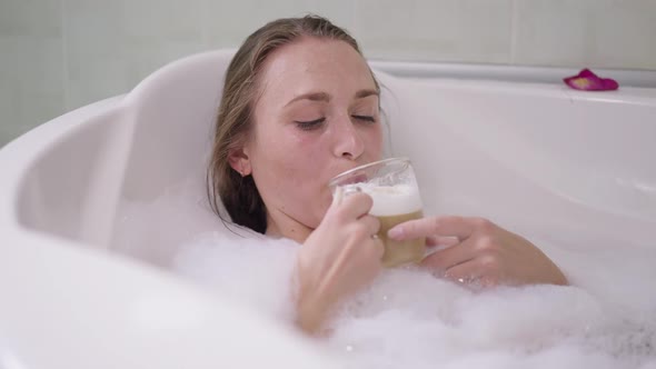 Happy Young Slim Woman Drinking Cappuccino Lying in Bath with Foam at Home