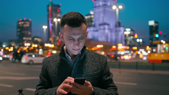 Business Man Is Using Phone App in City at Night with Blurred Cityscape Lights