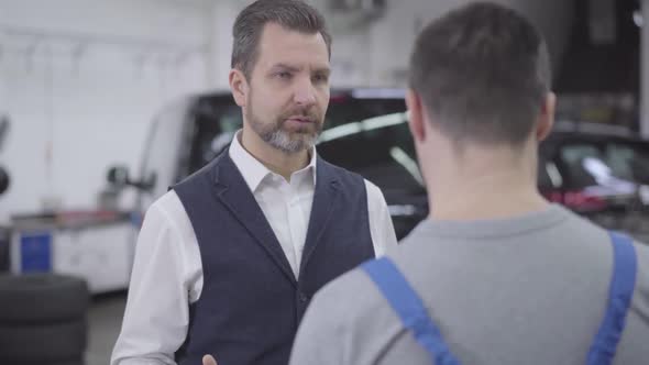 Close-up Portrait of Displeased Caucasian Man Talking with Unrecognizable Auto Mechanic. Adult