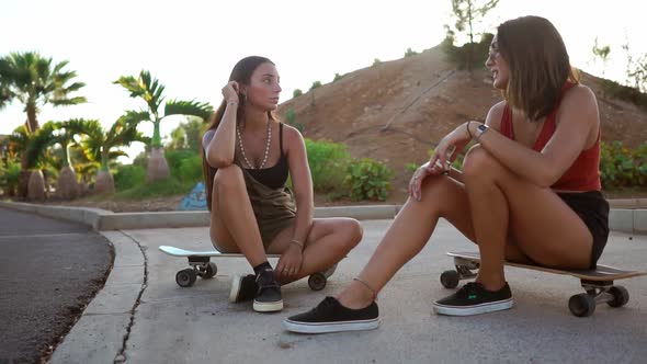Two Girlfriends Communicate in the Park Sitting on Skateboards Laughing and Smiling