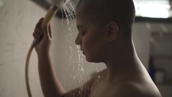 Sick and weak bald Asian girl is bathing with hand shower in bathroom is very sad and crying due to