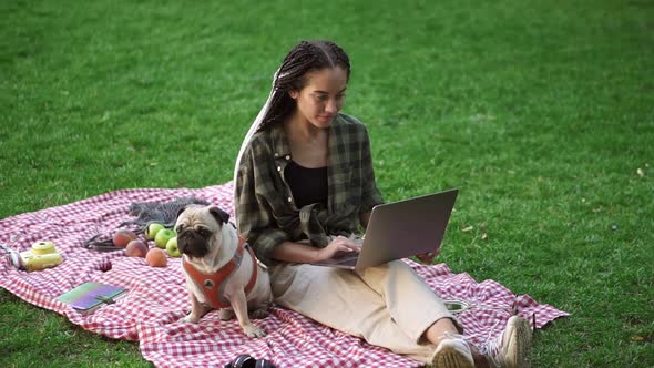Stylish Girl Sitting Outdoors on the Grass Lawn with Her Laptop Computer and Cute Little Pug is