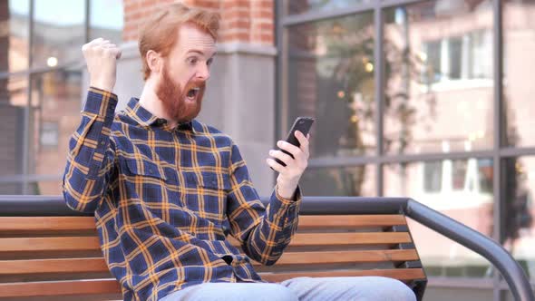 Redhead Beard Young Man Celebrating Win on Smartphone Sitting Outdoor on Bench