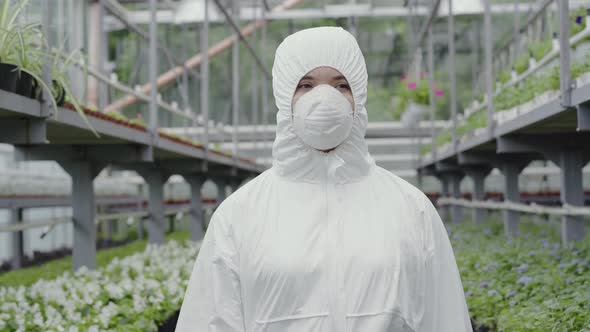 Confident Woman in Respirator and White Protective Workrobe Walking in Greenhouse. Portrait of