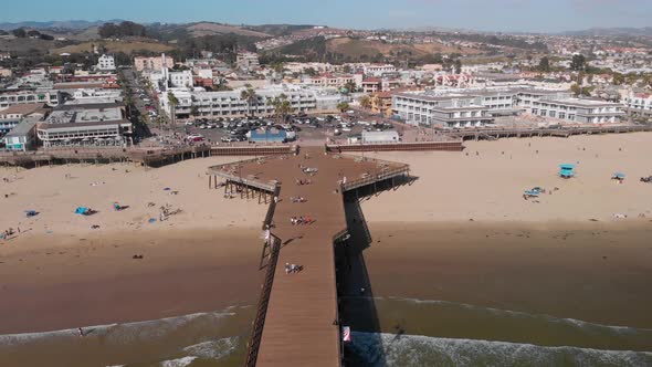 People walk and surf around Pismo Beach Pier as Drone Tilts Up Revealing Town