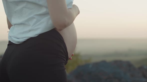 Confident White Pregnant Woman Stands Outdoor Holds Her Stomach and Raises Her Hands Up Side View