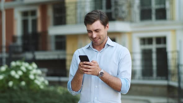 Happy Man Typing on Phone Outdoors. Man Writing Message on Phone at Street