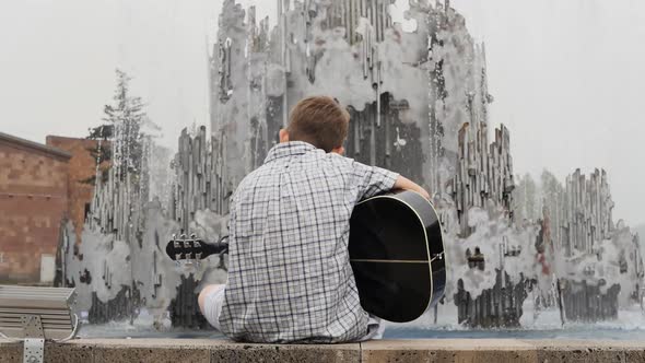 Teenage Boy in Shirt Playing Acoustic Guitar Facing Water Fountain in Public Park Sitting on