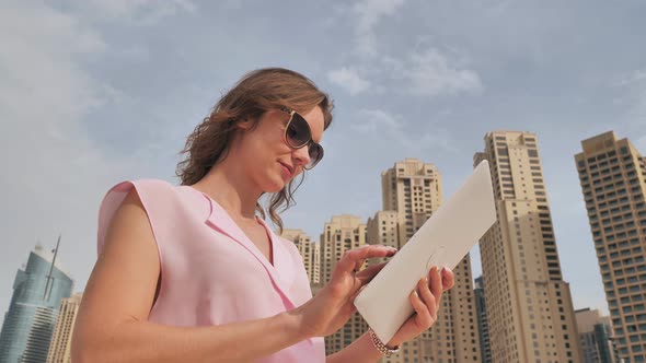 A Girl of European Appearance Works with a Tablet on the Background of Dubai Skyscrapers