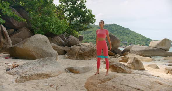 Woman Exercising on the Beach She Makes Rubber Band Squats Dressed in Pink Sportswear