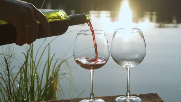 Red Wine Pouring From Bottle to Two Clear Glass on the Background of the Water Surface of the Pond