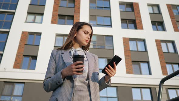 Woman in a Business Suit Looks at the Phone While Holding a Plastic Cup on the Background of a