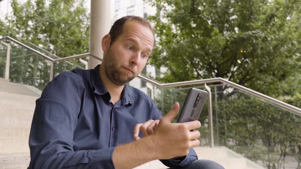 A Caucasian Man Takes Selfies on a Smartphone As He Sits on a Staircase in an Urban Area