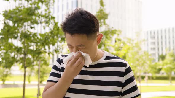 A Young Asian Man Blows His Nose with a Paper Tissue in an Urban Area  a Park and Office Buildings