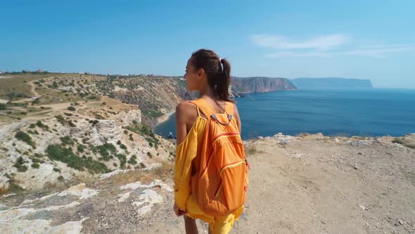 Rear View of Tanned Traveler Woman Standing on Cliff Edge, Posing in Bright Yellow Dress Blowing in