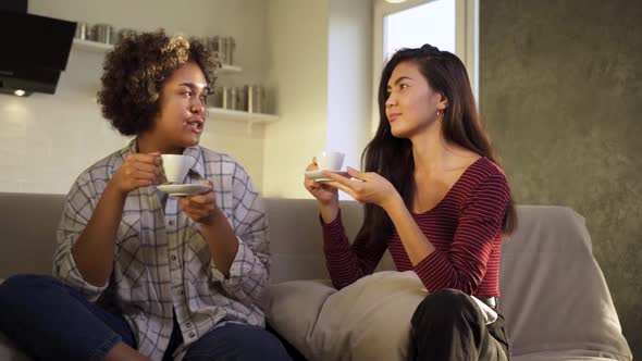 Two AfricanAmerican and Asian Girlfriends Laugh While Having Tea Together at Home