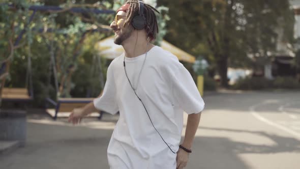 Relaxed Tattooed Caucasian Young Man with Dreadlocks Walking Along Sunny Urban Street Dancing To