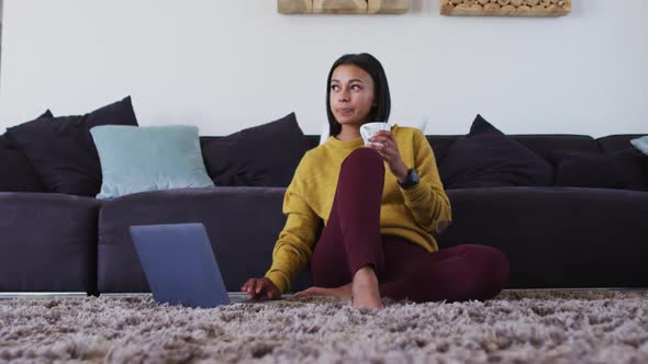 Mixed race woman sitting on floor using laptop drinking cup of coffee