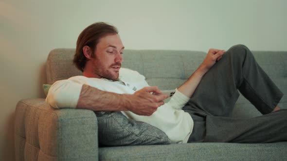 Positive long-haired man watching video on phone while lying on the sofa