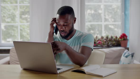 AfricanAmerican Ethnicity Man Freelancer Talking on Smartphone and Working on Laptop at Home