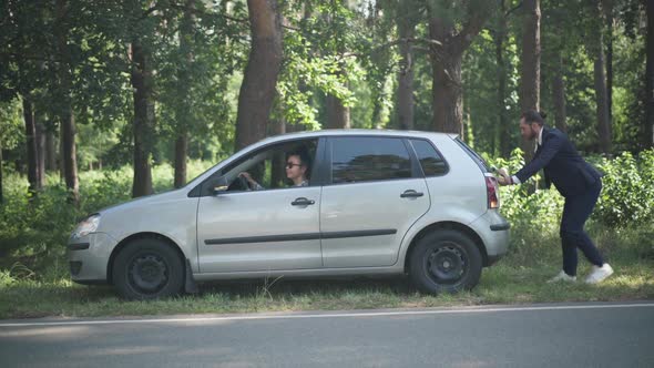 Young Caucasian Woman Sitting on Driver's Seat As Man Pushing Vehicle Outdoors on Sunny Day