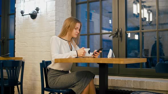 Young Business Woman Is Using Smartphone App and Drinking Coffee in the Cafe