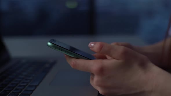 Closeup Hands of Young Woman Scrolling Mobile Phone Sitting at Desk in Dark Office Room