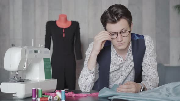 Portrait of Puzzled Man Sitting at the Table in Atelier and Thinking. Brunette Caucasian Male Tailor