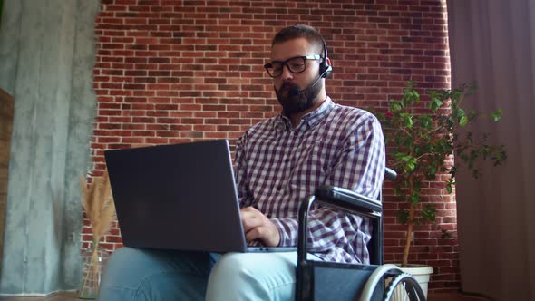 Man in Glasses is Sitting on Wheelchair and Working on Laptop with Headset