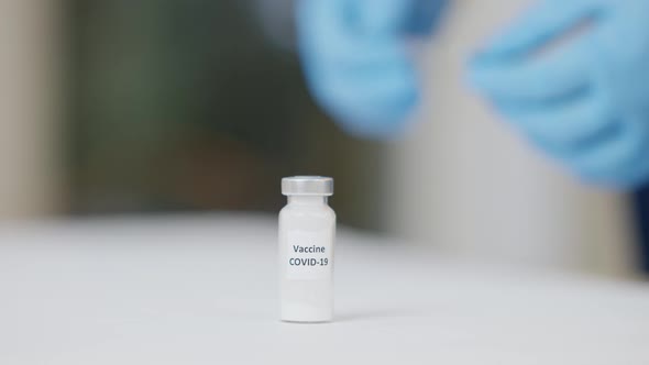 Closeup of Coronavirus Vaccine Ampoule with Blurred Hands in Protective Gloves Putting Syringe on