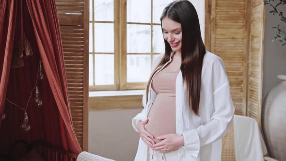 Happy Pregnant Woman Strokes Big Belly Standing Near Crib