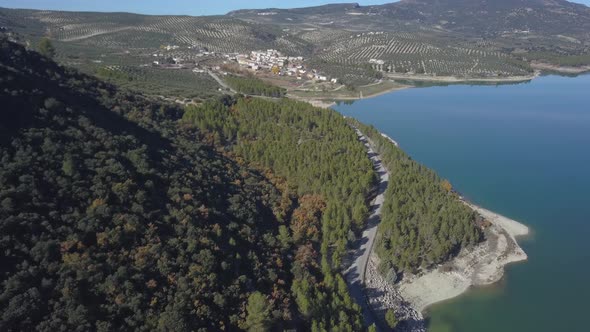 Aerial view of a road surrounded by a forest close to a lake with a small village in the horizon. Sp